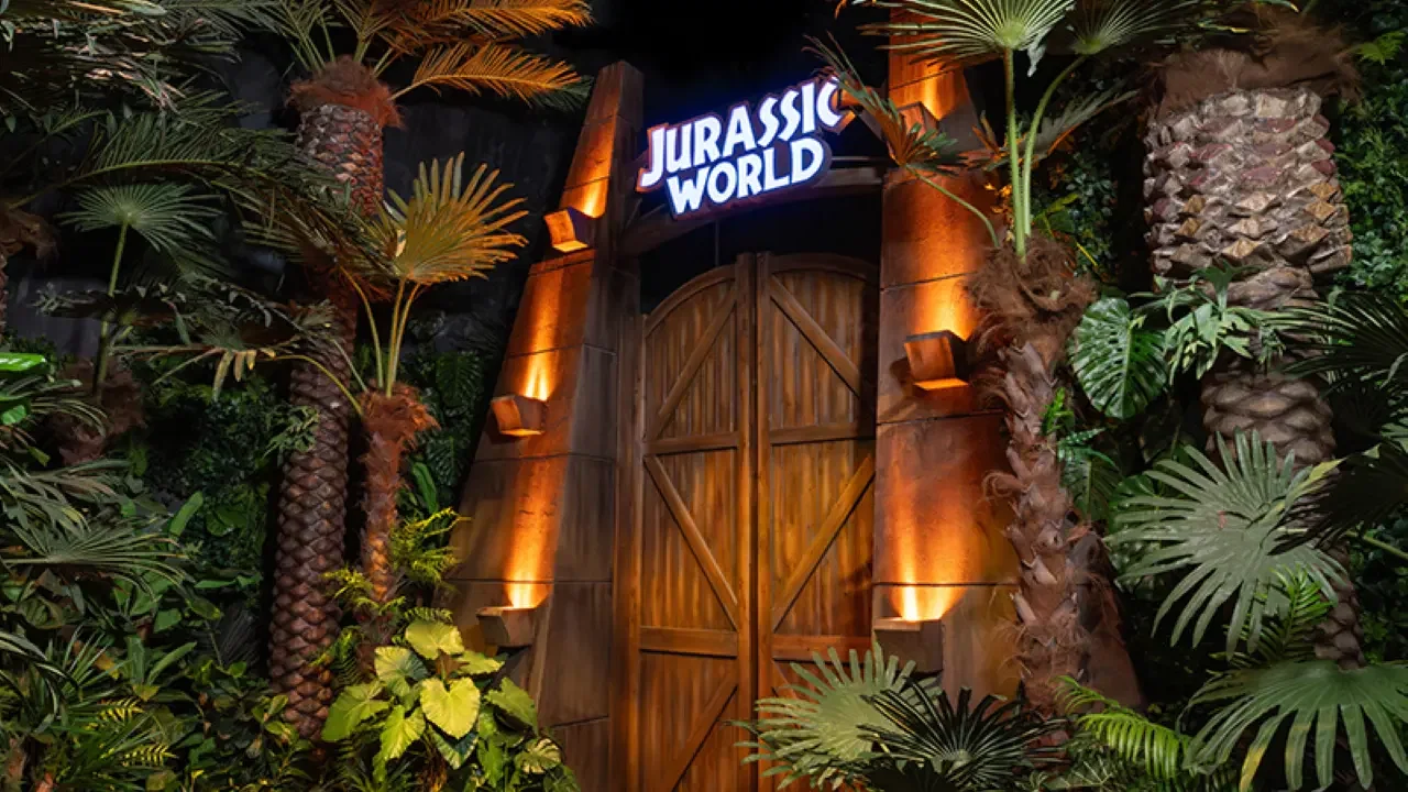Closed wooden double doors featuring lit wall sconces and a glowing park logo overhead are framed by dense tropical palm trees and foliage at Jurassic World: The Experience Bangkok Ticket.