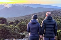 Two hikers in down jackets holding hands and looking out over a vast mountain landscape at sunrise.