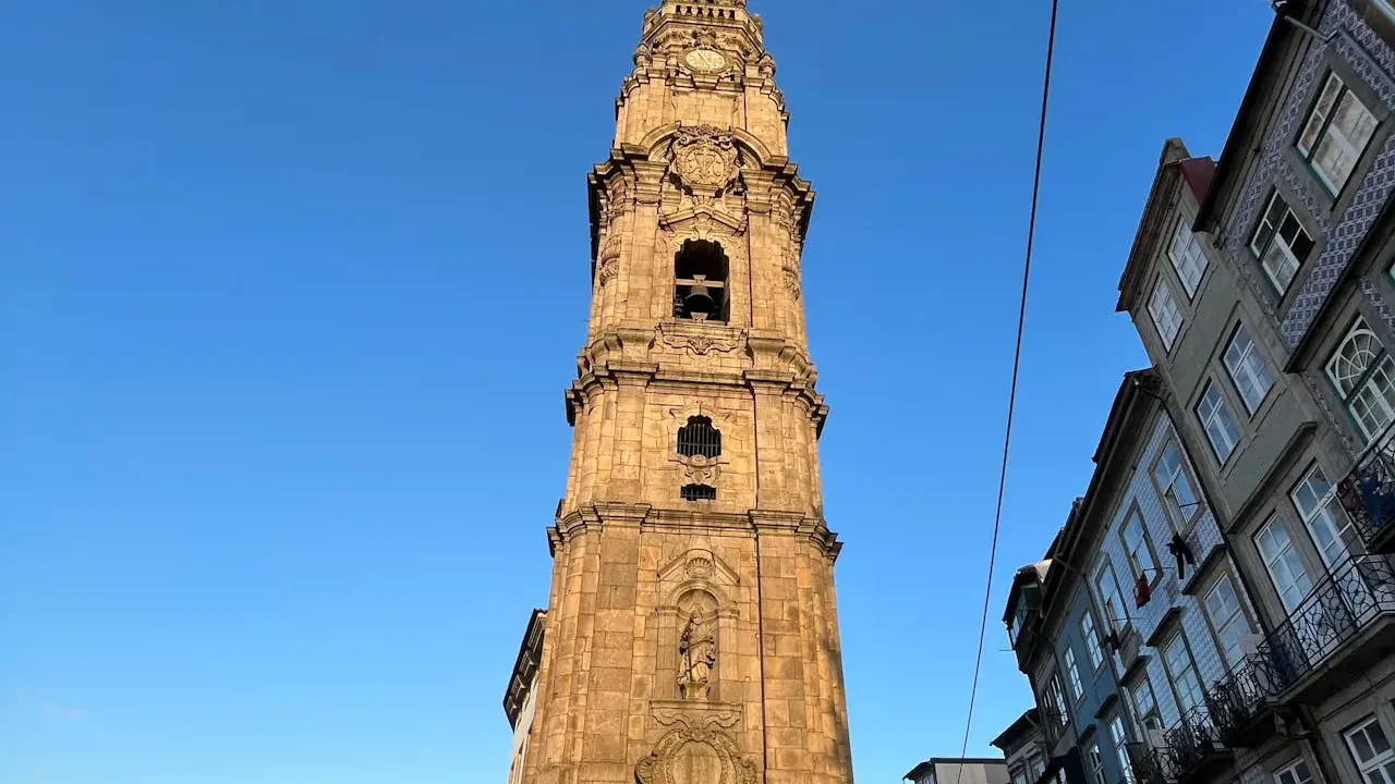 A tall stone bell tower stands beside traditional multi-story buildings with balconies under a blue sky at Clérigos Tower & Museum Ticket.