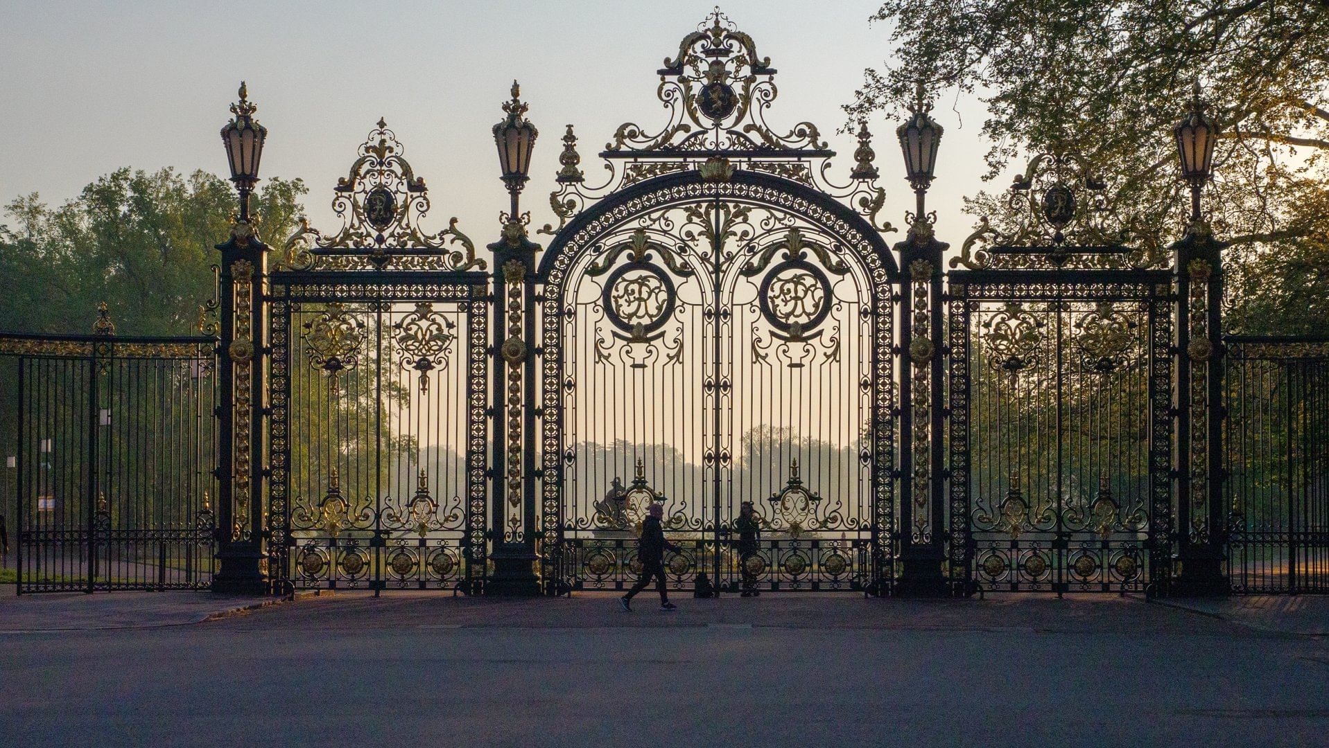 The ornate wrought-iron entrance gates of Parc de la Tête d'Or in Lyon, with intricate details and a silhouette of people walking through at