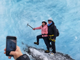 A person is taking a photo on it's phone of a couple standing by an ice wall on the glacier