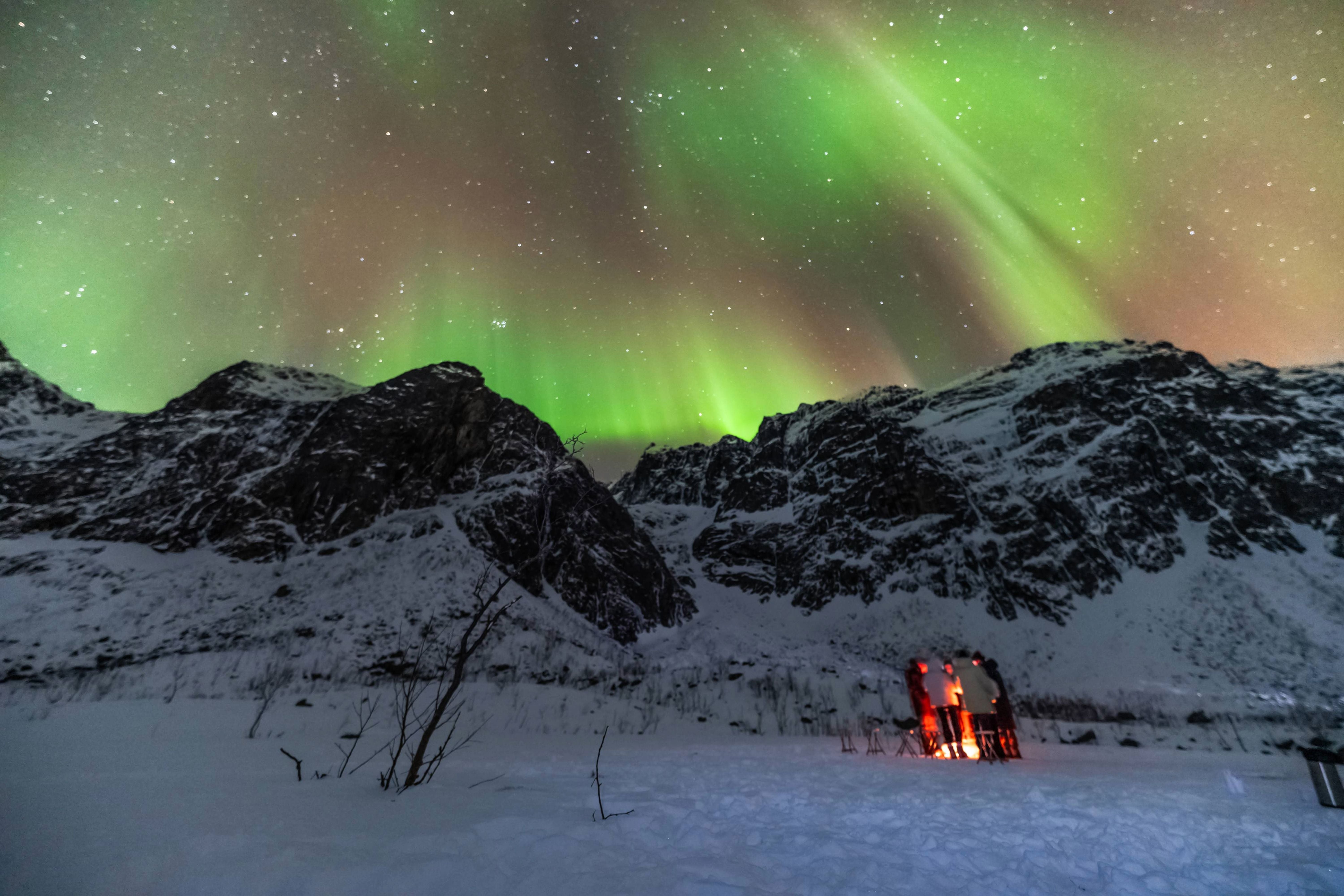 Glowing campfire burning on snow in front of majestic Arctic mountains under the green Northern Lights.