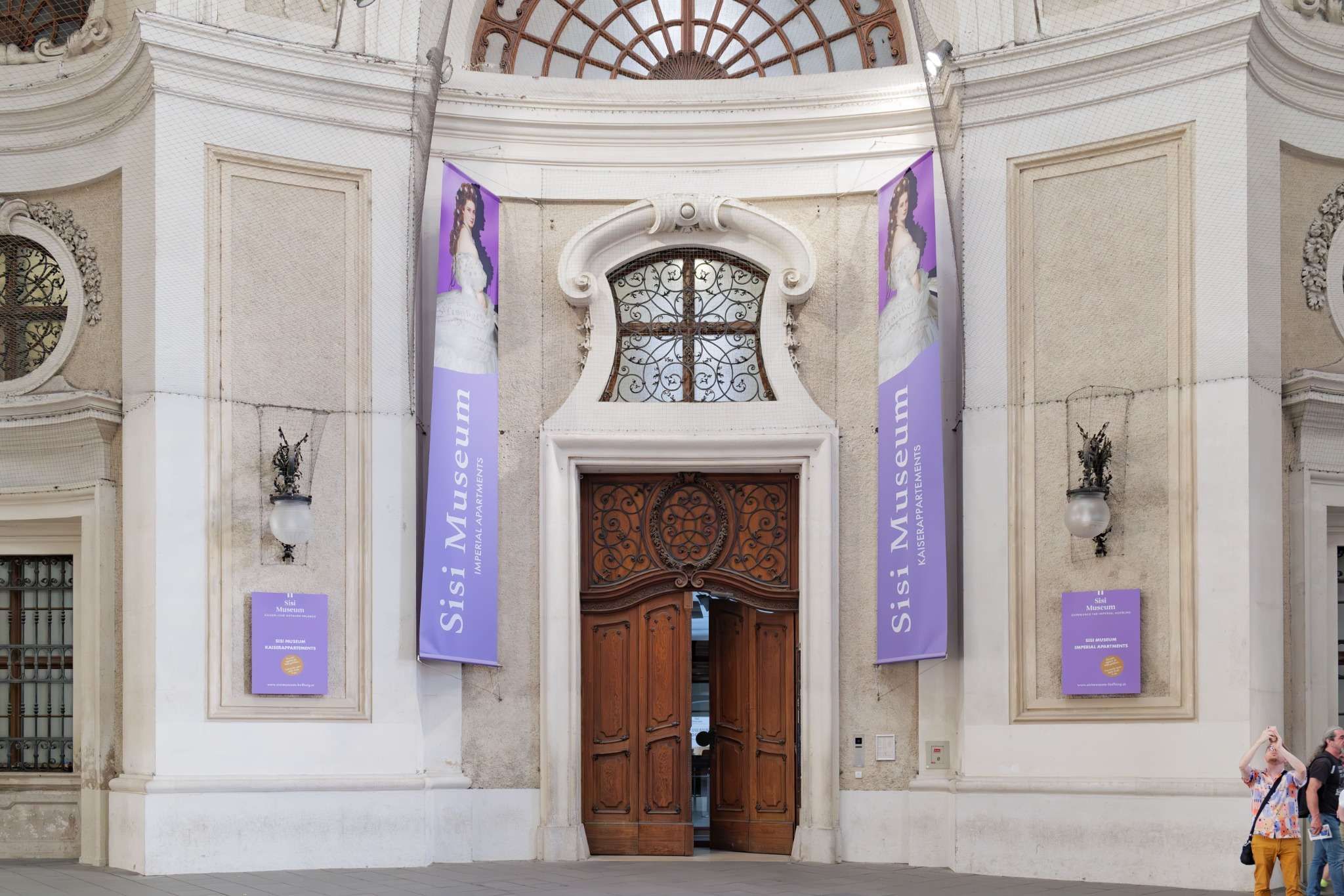 Ornate arched doorway with wooden double doors, flanked by purple Sisi Museum banners and signs inside the Hofburg Palace