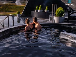 Two people bathing in a circular geothermal pool at Krauma, facing away toward the steaming landscape and open West Iceland plains, with the