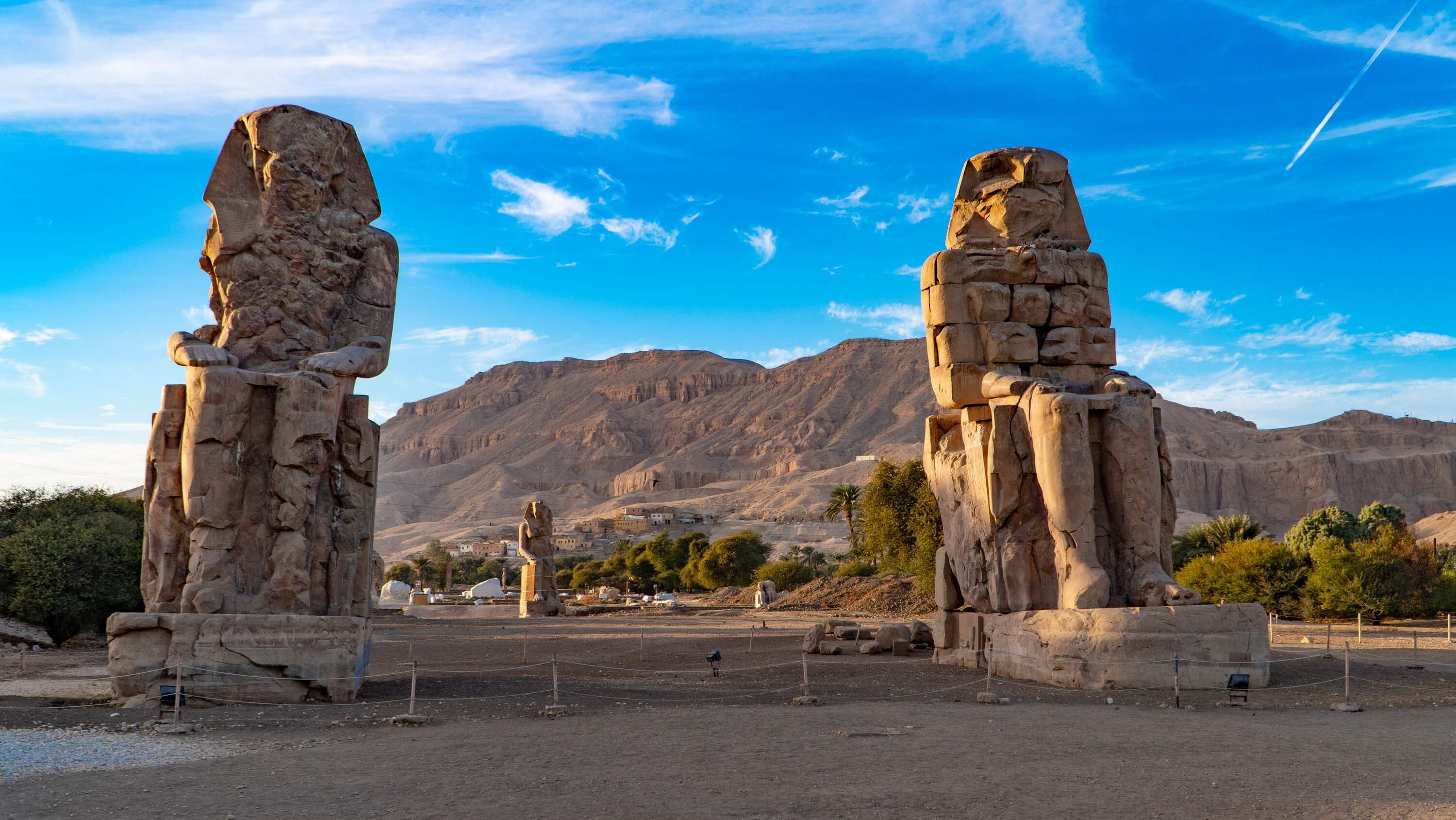 Two giant ancient stone statues, known as the Colossi of Memnon, standing in an open area with mountains and a blue sky in the background ne