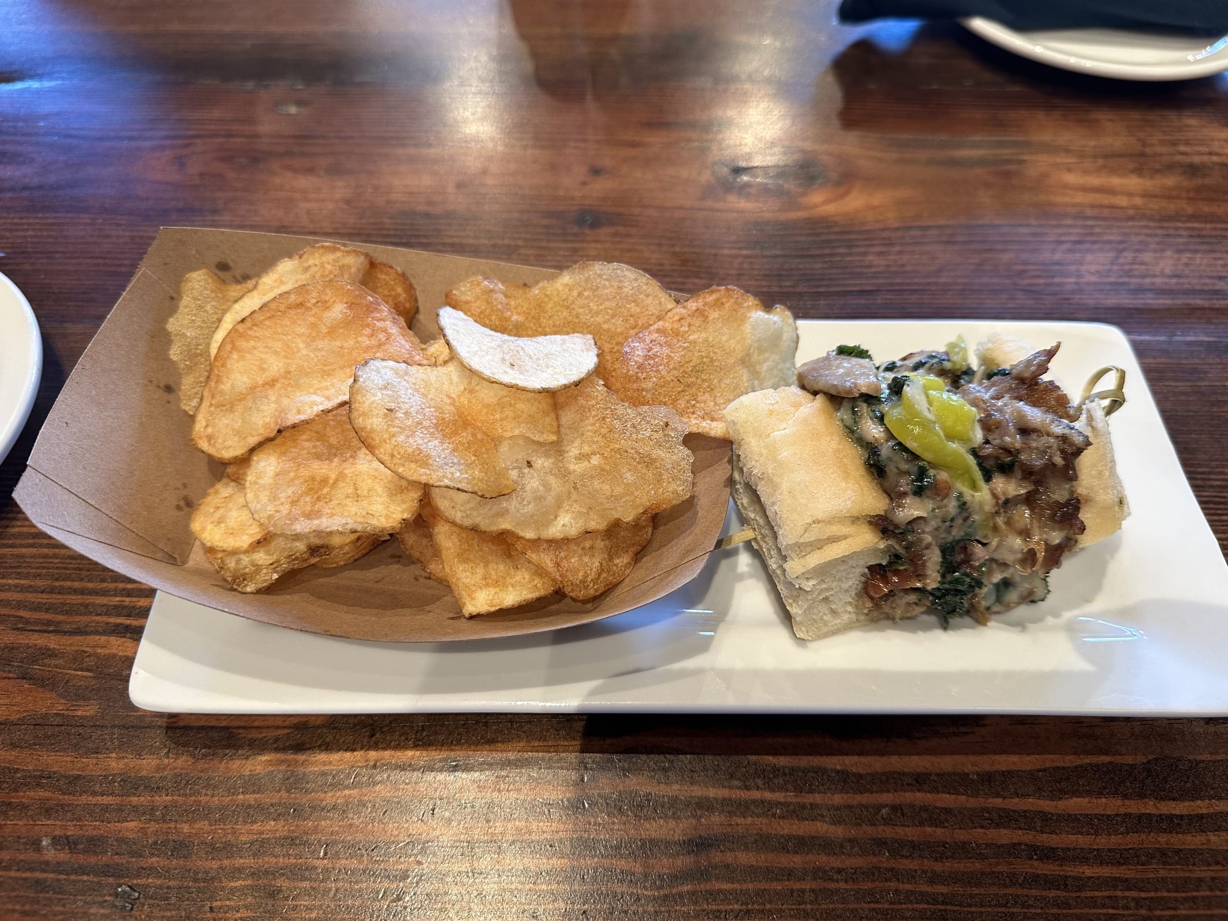 A porchetta sandwich on a sub roll with greens, melted cheese, and a banana pepper, served with house-made potato chips on a wooden table at Dressel's Public House