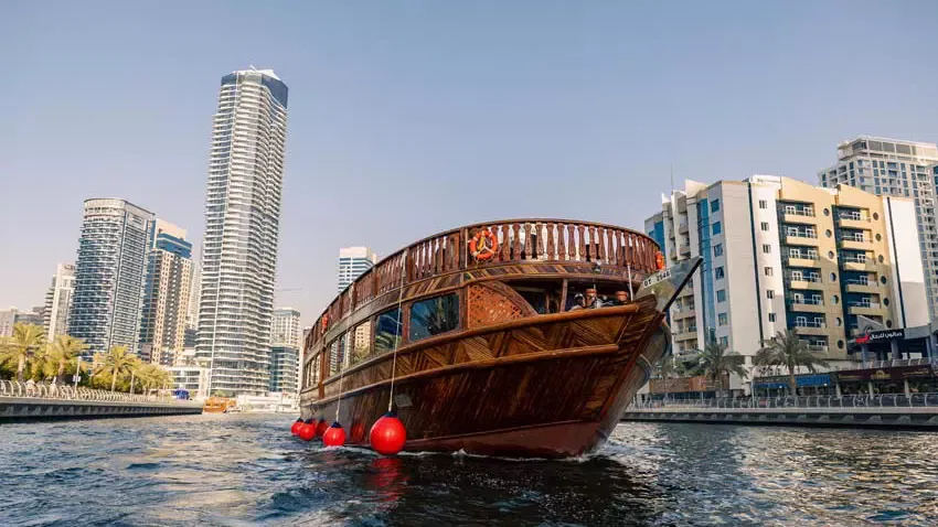 A wooden dhow boat cruises past towering skyscrapers lining the waterfront at Alexandra Dhow Cruise in Dubai Marina.