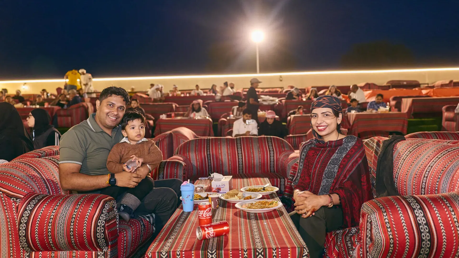 A family sits together at a long table with food and drinks in an outdoor seating area at Desert Safari + Al Faqaa Camp.