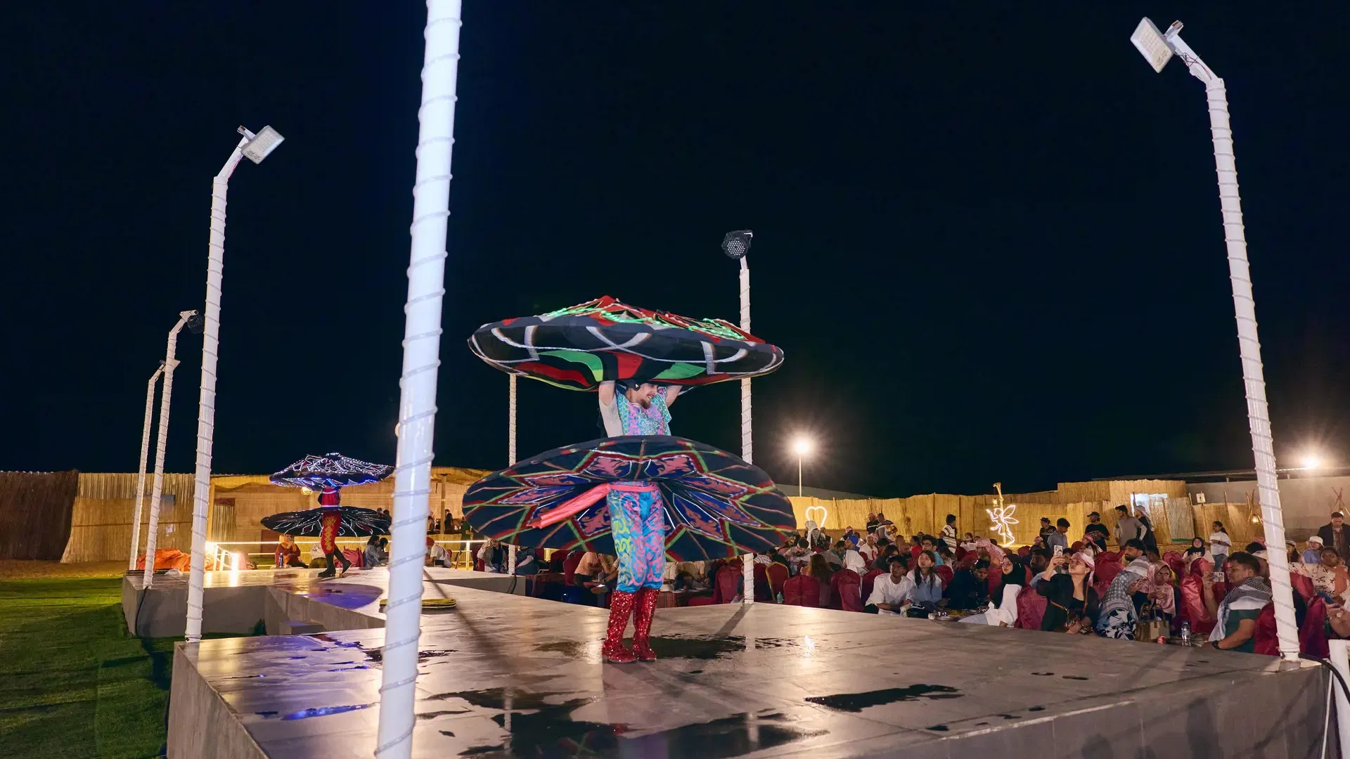 A performer in a vibrant spinning costume performs a dance on stage in front of an audience at Desert Safari + Al Faqaa Camp.