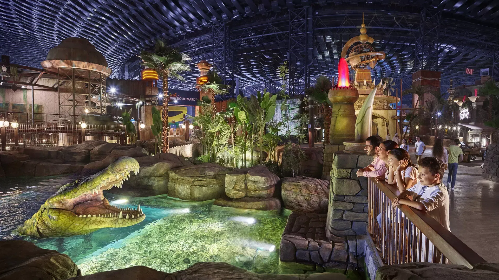 A family leans on a railing overlooking an artificial pool with a mechanical crocodile while other visitors walk around at IMG Worlds of Adventure Theme Park.