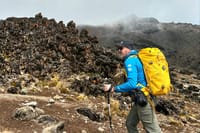 A hiker with a yellow backpack and trekking poles walking past dark volcanic rock formations on a misty mountain trail.