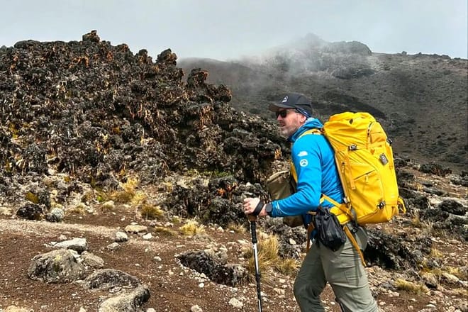 A hiker with a yellow backpack and trekking poles walking past dark volcanic rock formations on a misty mountain trail.