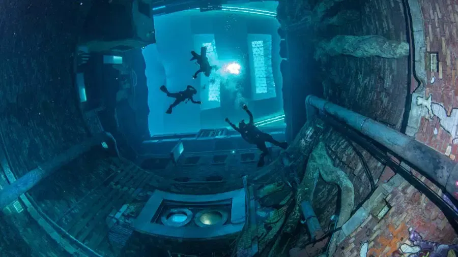 Divers explore the depths of a sunken town featuring brick walls and pipes with light filtering down from above at Deep Dive Dubai.