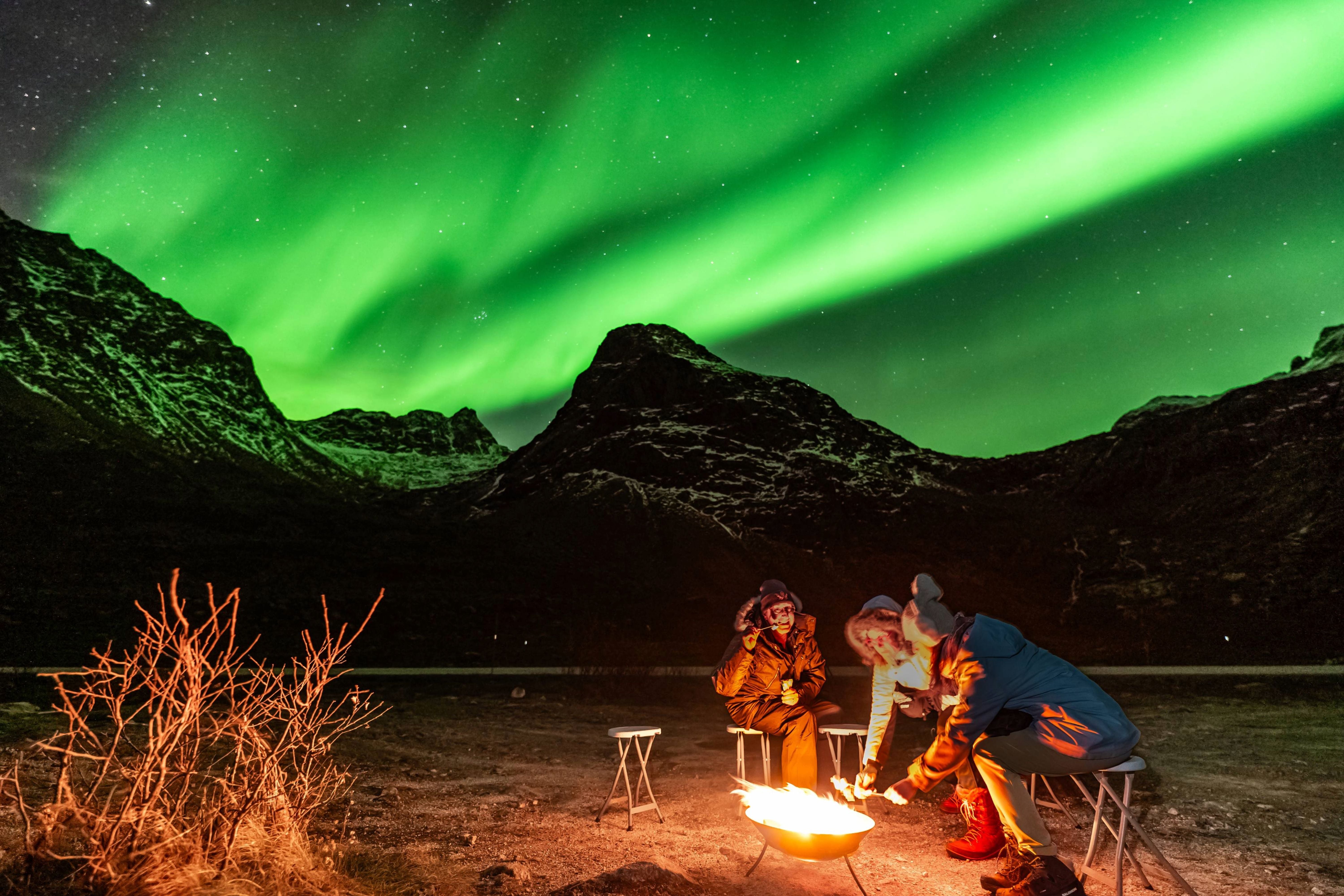 Small group of travelers sitting on stools around a glowing campfire in a snowy mountain valley beneath a bright green Northern Lights displ