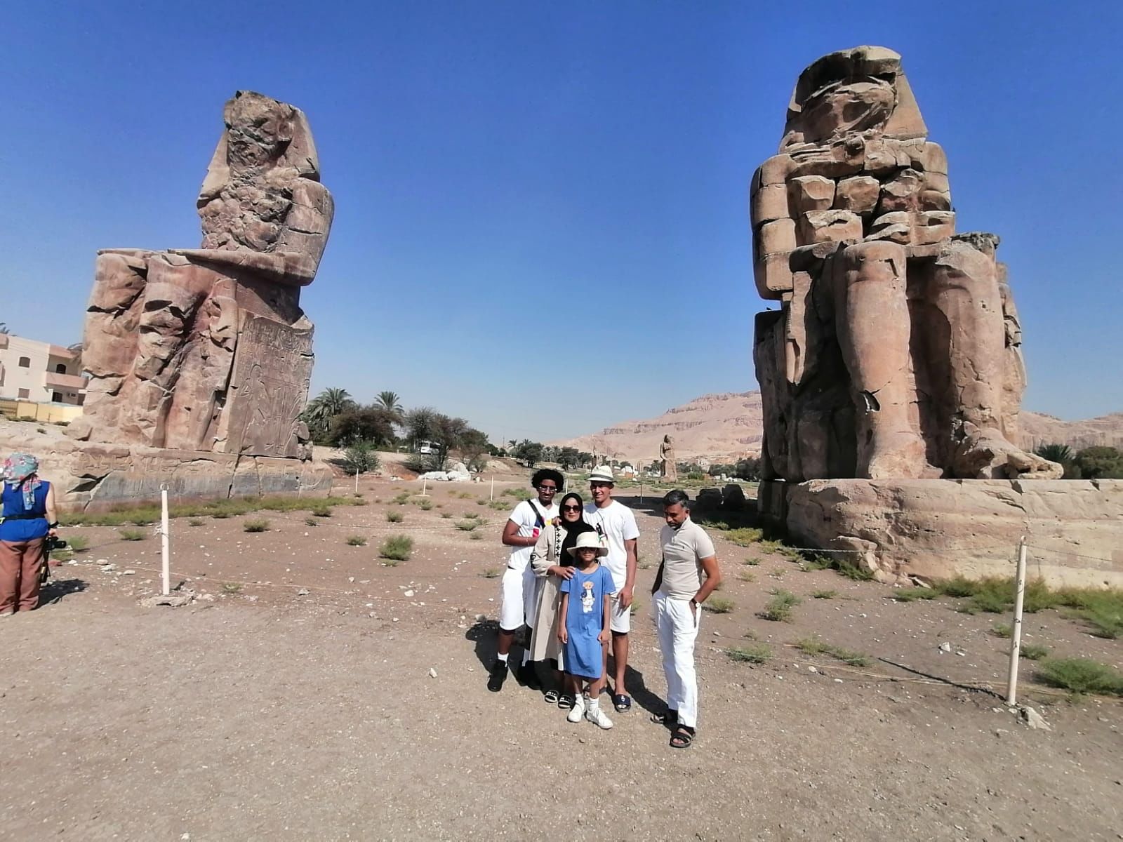 Par une journée ensoleillée, une famille se tient debout devant les immenses statues des colosses de Memnon, avec un ciel bleu clair et un paysage désertique dans le
