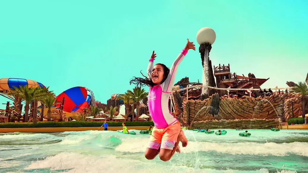 A young girl with her arms raised jumps happily in the wave pool with other visitors floating on inner tubes at Yas Waterworld, Abu Dhabi.