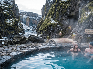 Couple soaking in the steaming geothermal pool at Húsafell Canyon Baths with mossy lava canyon walls rising behind them