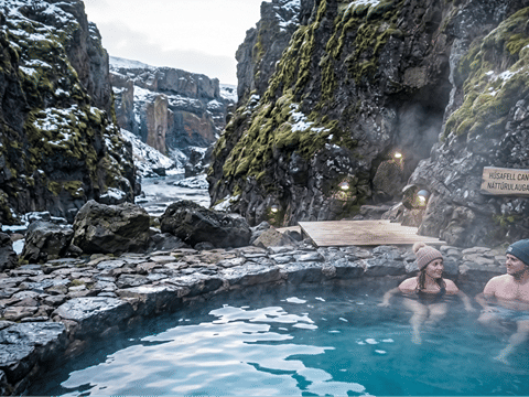 Couple soaking in the steaming geothermal pool at Húsafell Canyon Baths with mossy lava canyon walls rising behind them