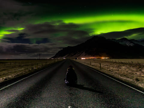 Girl sits on road in Iceland looking at the Northern Lights