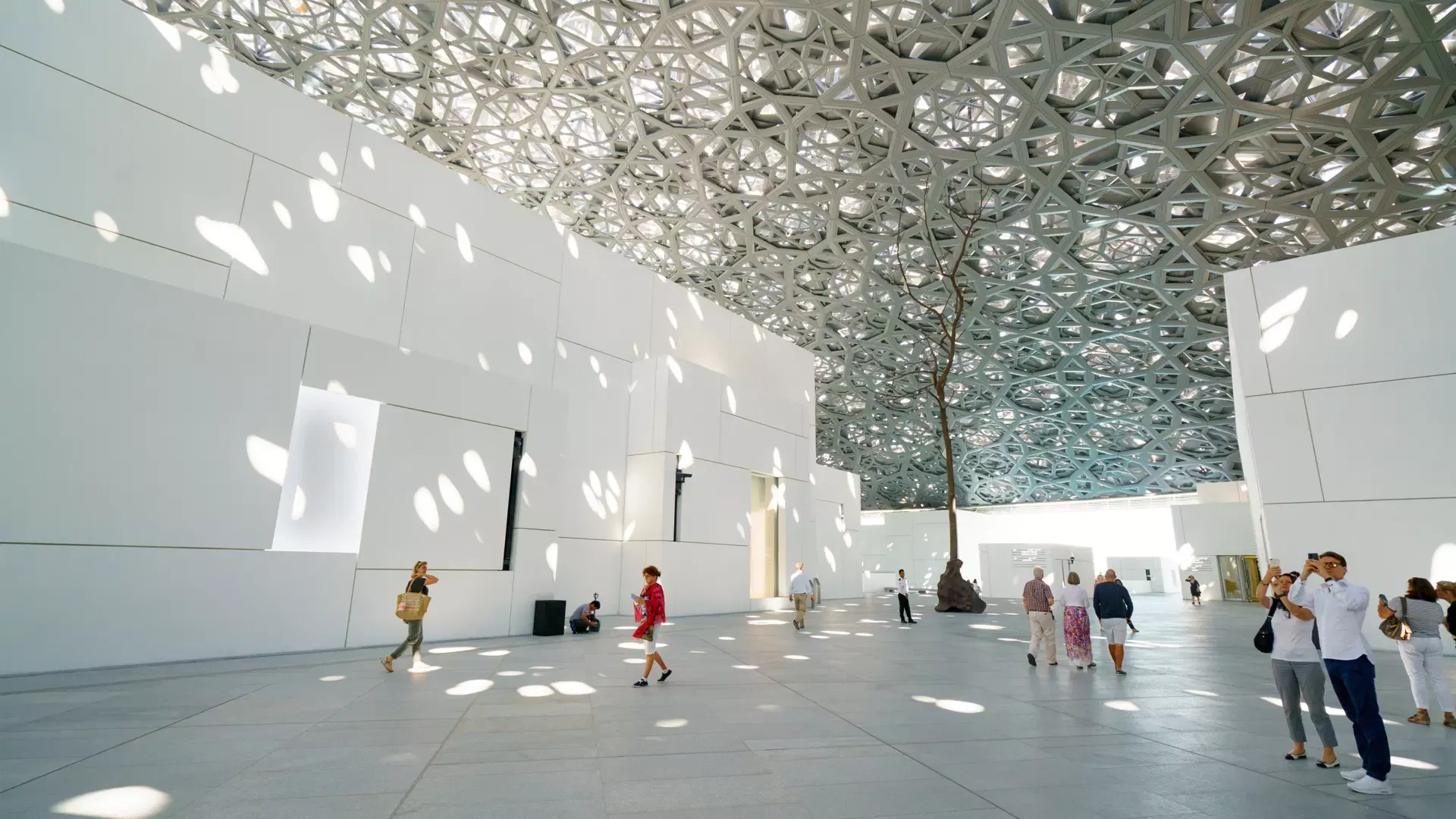 Visitors crossing a bright white plaza as dappled sunlight falls through the latticed dome onto the walls and floor at Louvre Abu Dhabi