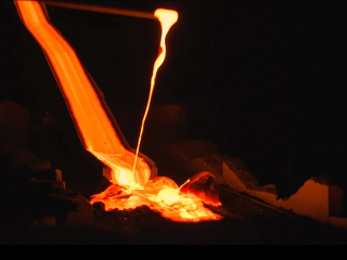 Molten lava flows in bright orange from a tilted container into a cooling pit, glowing intensely against a dark background.