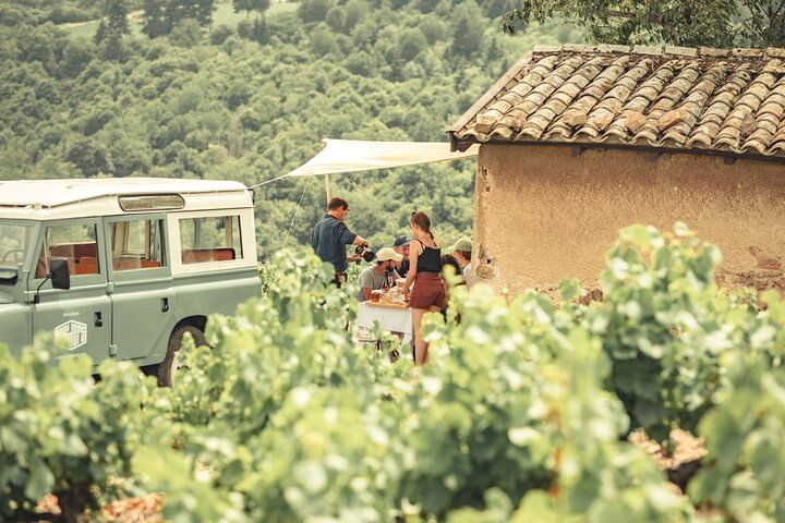 By a small hut in a vineyard, a group of people are enjoying a picnic. It's a sunny summer's day. A vintage Land Rover is parked nearby.