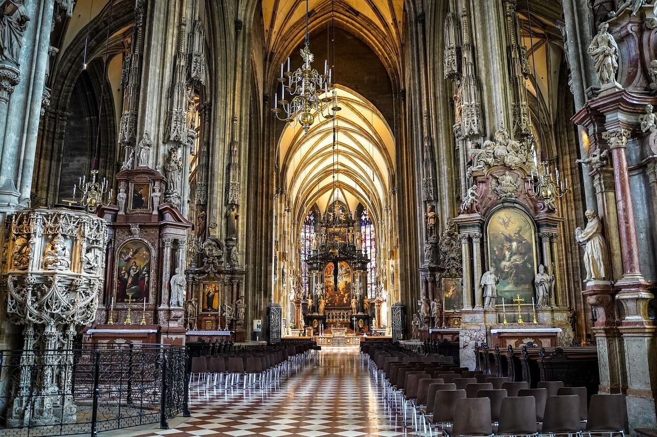 View down the central aisle of St. Stephen’s Cathedral, with vaulted ceilings, ornate altars, religious paintings, and rows of pews.