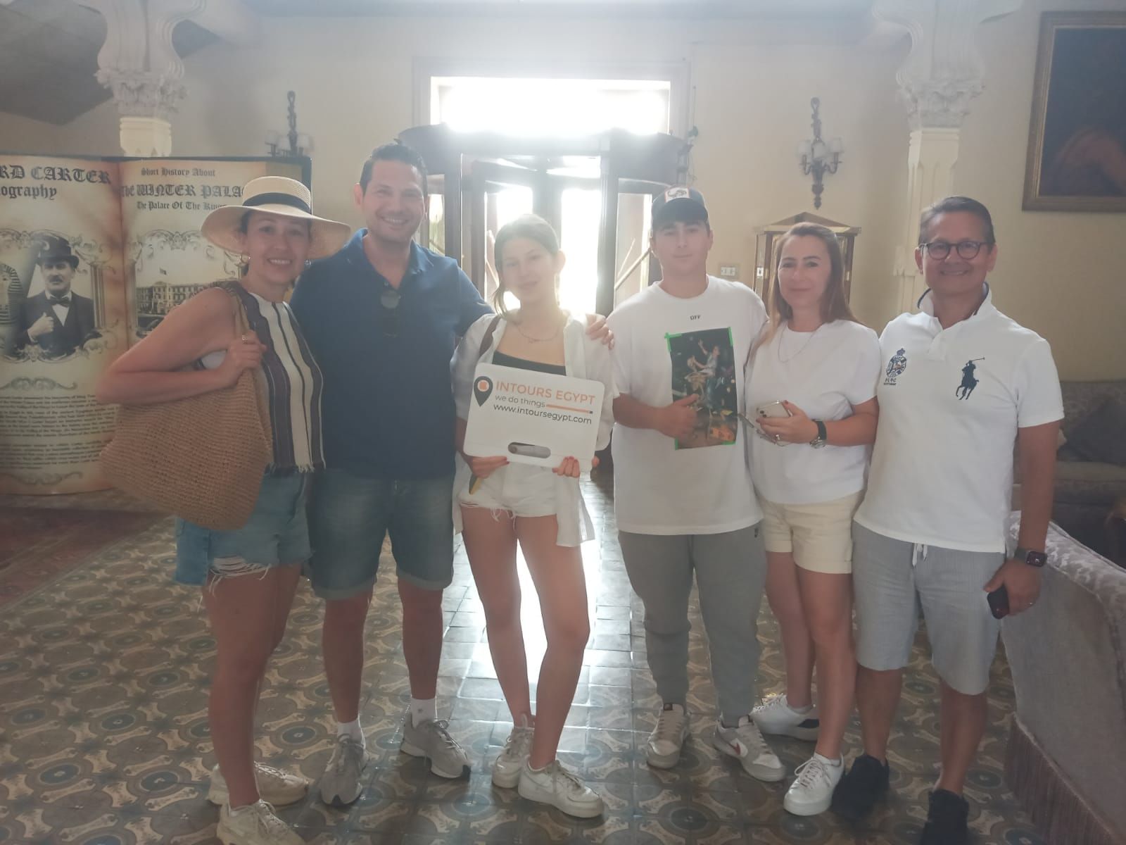 Six personnes se tiennent debout ensemble à l'intérieur, souriantes pour une photo de groupe ; une personne tient une pancarte Intours Egypt, entourée d'une décoration vintage
