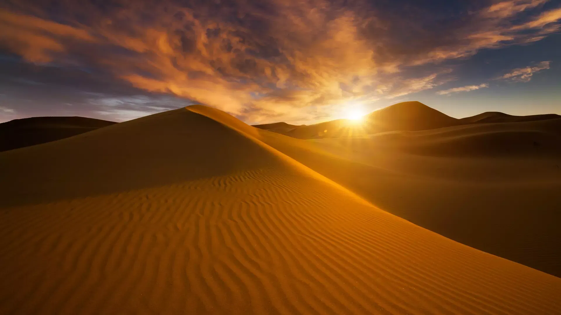 Golden sand dunes stretch under a dramatic sunset sky with vibrant orange clouds at Desert Safari + Al Faqaa Camp.