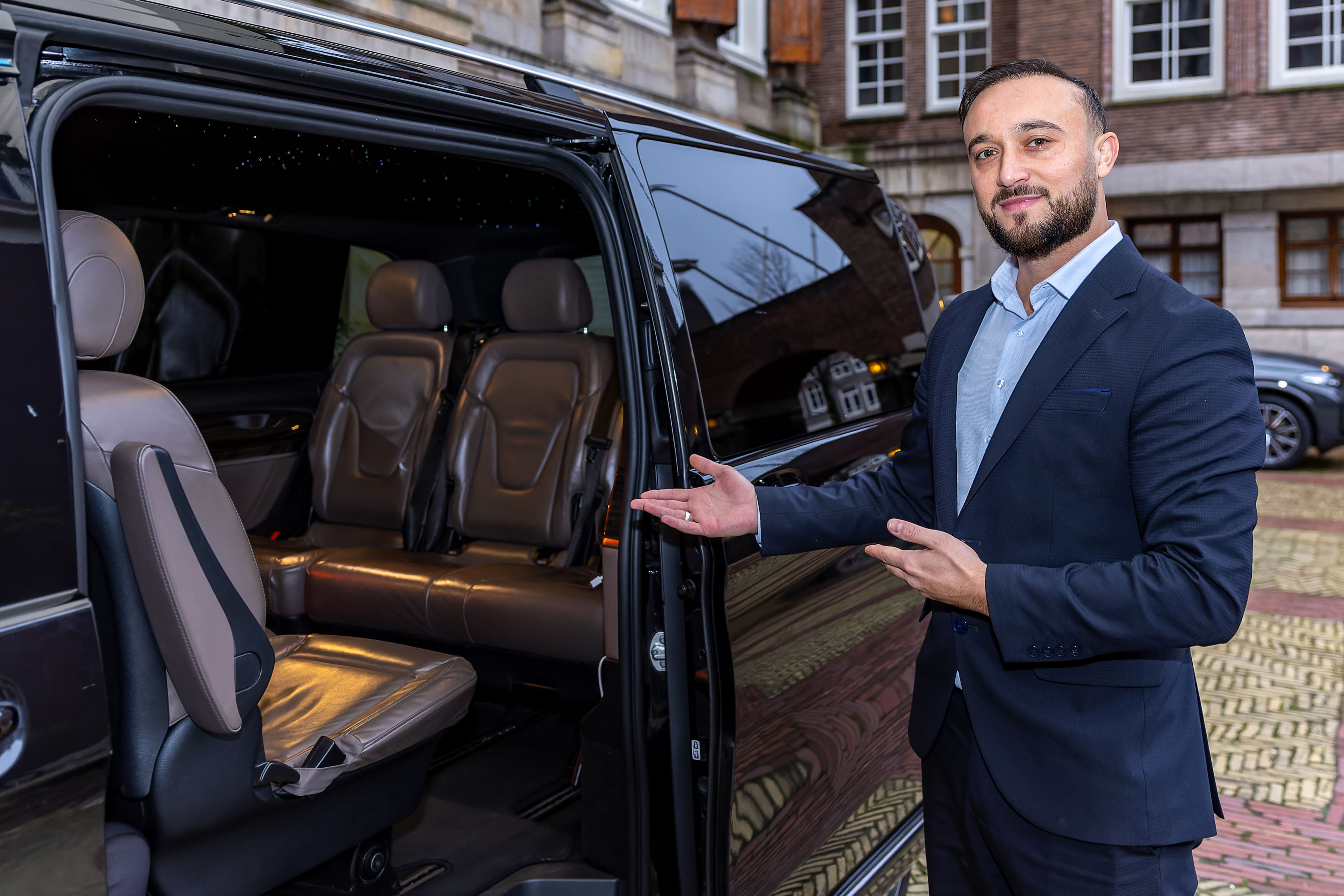 A local guide in a suit is standing next to an open luxury van with leather seats, gesturing toward the vehicle's interior.