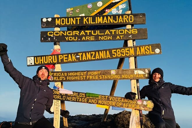 Two hikers celebrating at the Uhuru Peak summit sign on Mount Kilimanjaro, Tanzania, at an altitude of 5,895 meters.