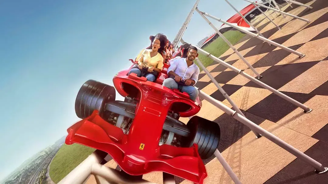 Two riders in a red Formula One-shaped coaster car climbing a steeply banked chequered track under a clear blue sky at Ferrari World Abu Dhabi