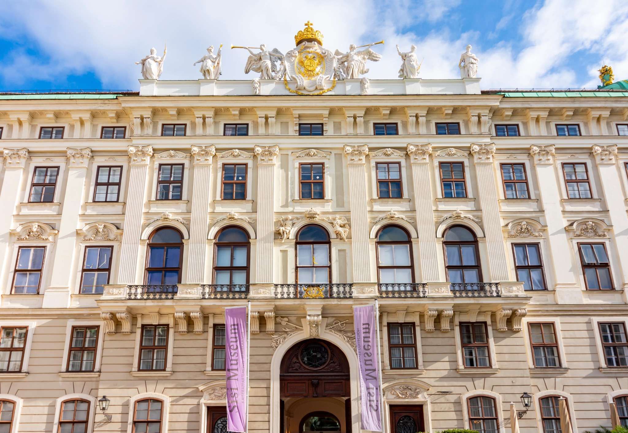 Grand white facade of the Hofburg Palace adorned with statues and a golden emblem, with purple Sisi Museum banners over the central entrance