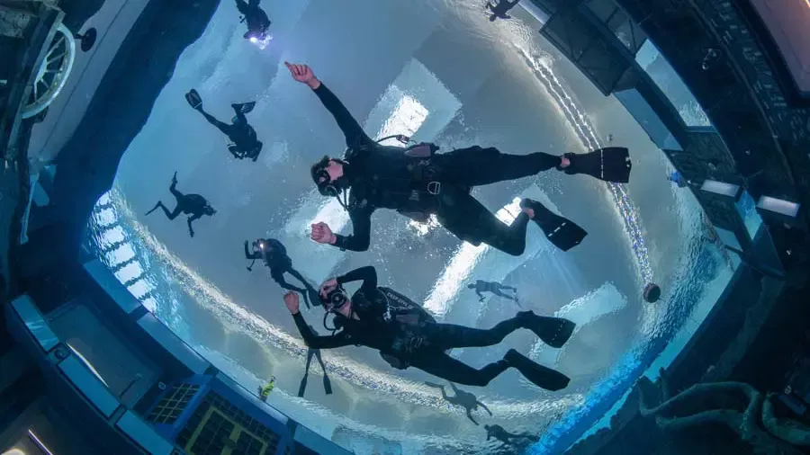 Multiple scuba divers swim vertically upwards inside a flooded urban set with a bright surface visible above at Deep Dive Dubai.