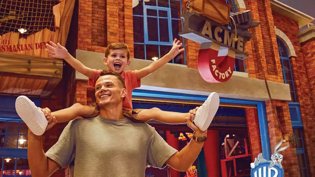 A smiling boy sits on a man's shoulders with arms raised in front of a brick building with an Acme Factory sign at Warner Bros. World Abu Dhabi Theme Park.