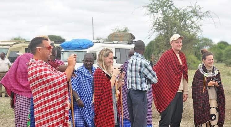 A group of tourists draped in red and patterned Maasai shúkà cloaks walking outdoors alongside local guides.