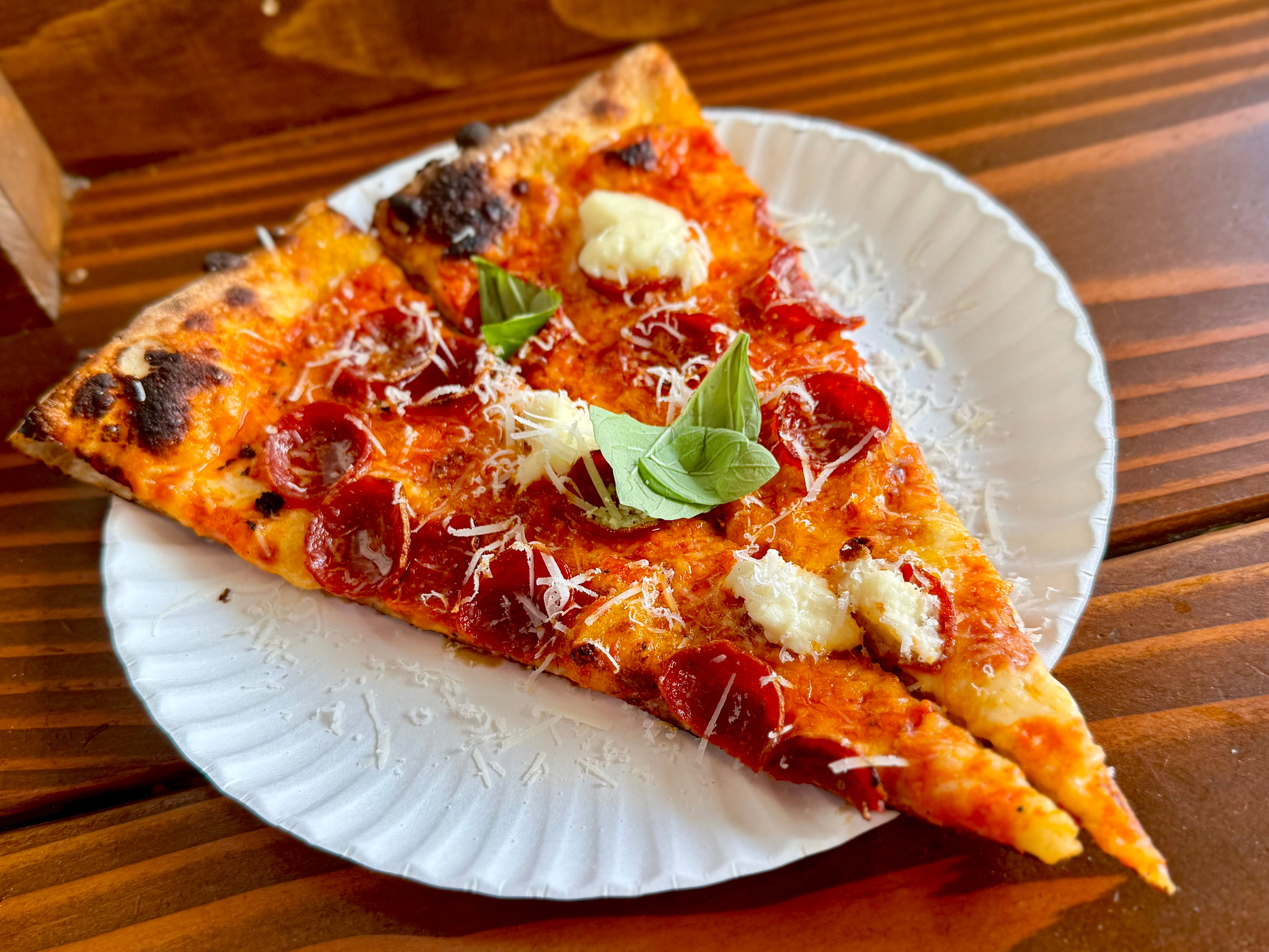 A pepperoni pizza slice with creamy sauce, cheese, and fresh basil on a paper plate.