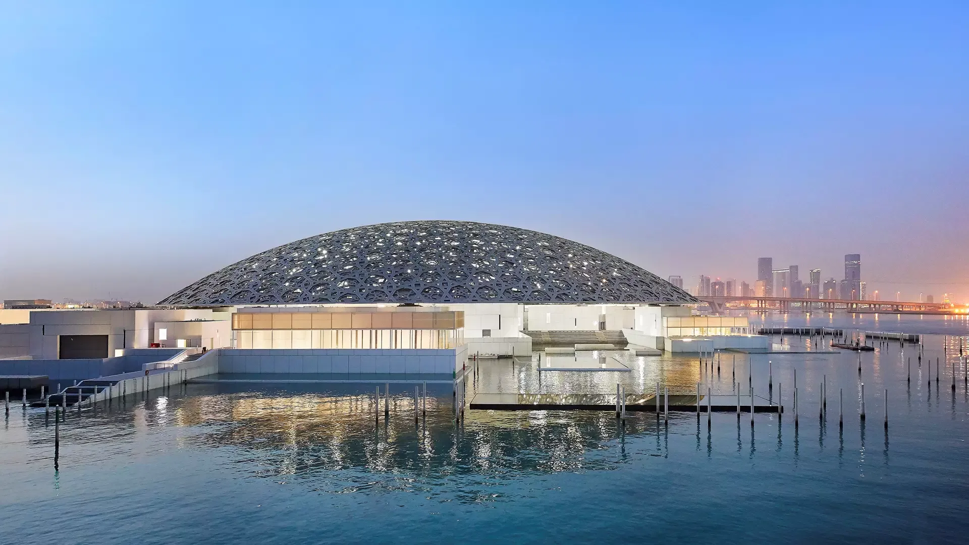 The full silvery dome rising above the water at twilight with the Abu Dhabi skyline on the horizon behind it at Louvre Abu Dhabi