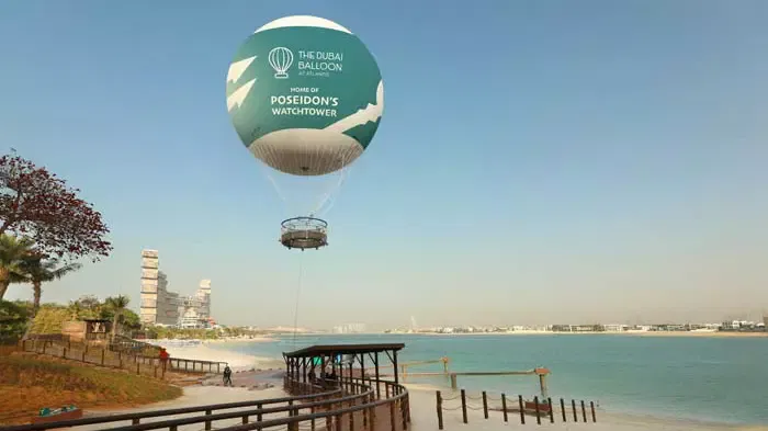 A large tethered gas balloon hovers above a boardwalk and water near a beach at The Dubai Balloon at Atlantis.