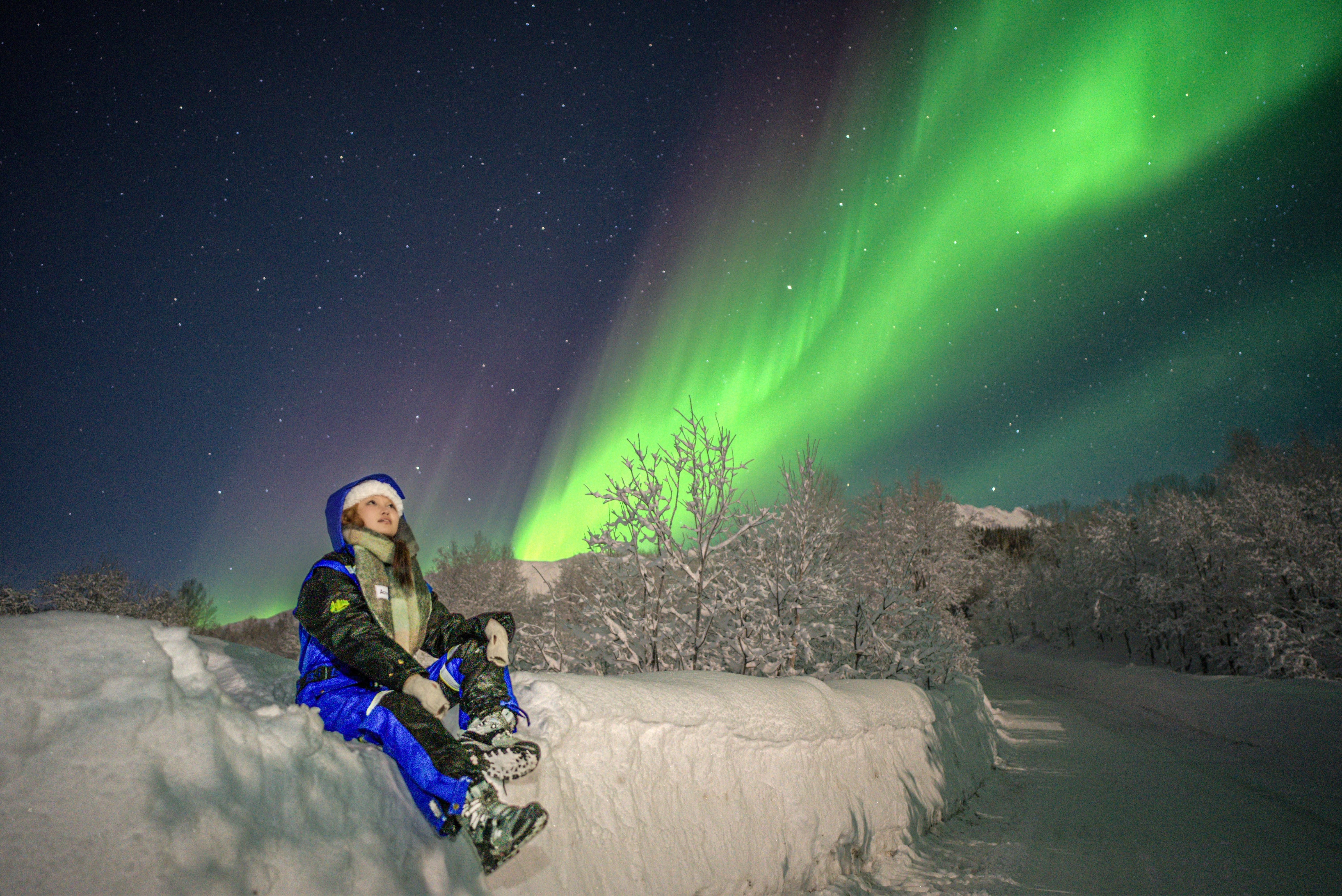 Traveler wearing a thick blue thermal suit sitting on a snowbank under a vibrant green and purple Northern Lights display.