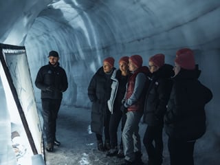 Specialist glacier guide addresses a small group of five visitors wearing red beanies inside a curved blue ice tunnel at Langjokull glacier