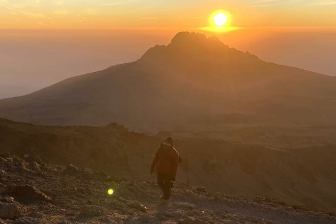 A silhouette of a hiker walking towards a golden sunrise behind a mountain peak on Mount Kilimanjaro.