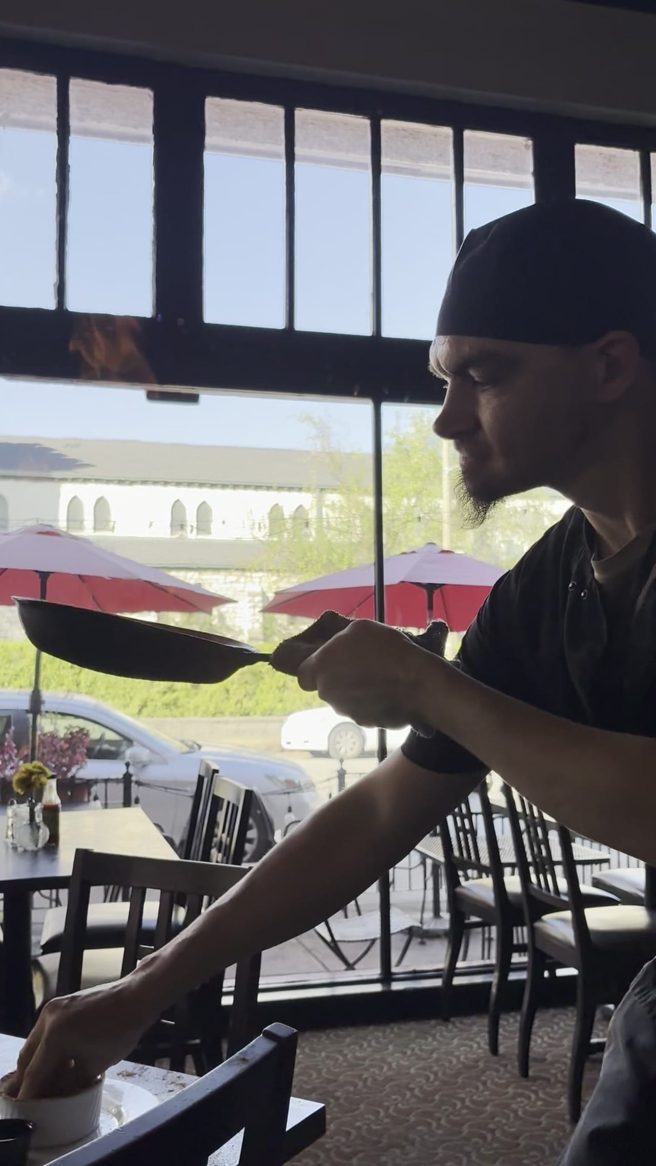 Silhouette of chef with pan inside restaurant, bright window with red sidewalk umbrellas visible outside