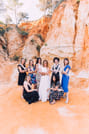 A candid group photo of several women laughing and standing together on a shoreline, with soft waves washing up on the sand in the backgroun