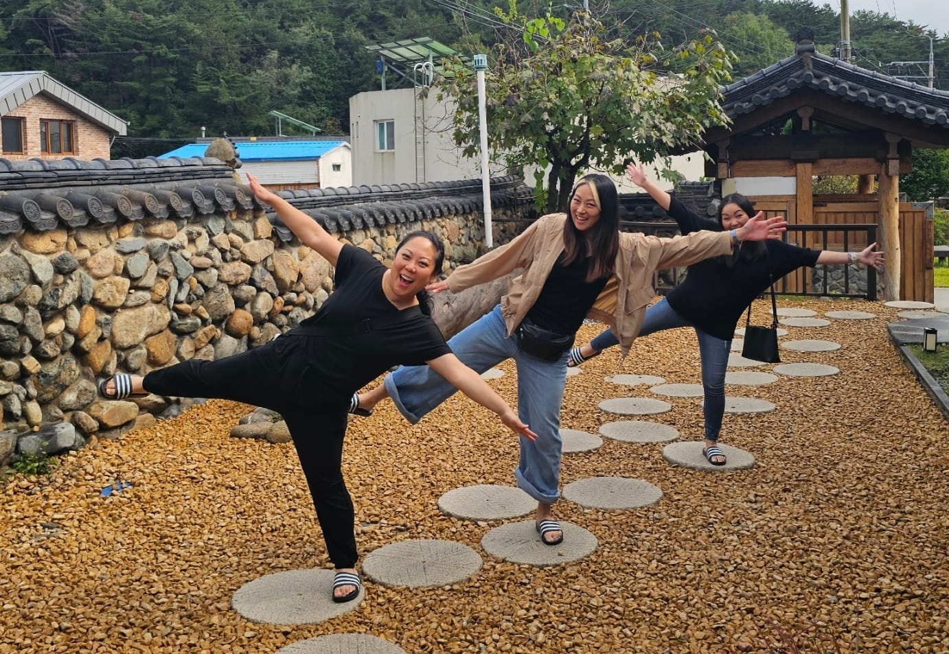 Three friends balancing on stepping stones and posing in the outdoor yard of a hanok café.