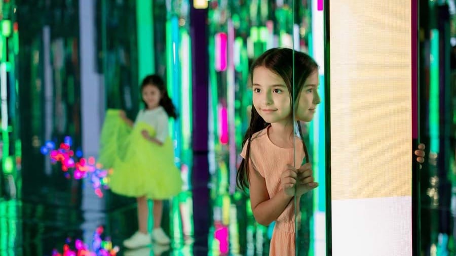 A girl standing between mirrored panels in a green-lit mirror maze at AYA Universe Dubai, with another child reflected behind her