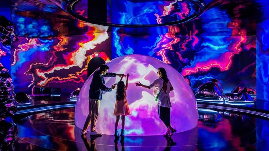 A family reaching towards a large glowing dome while swirling blue, pink and orange projections move across the surrounding walls at AYA Universe Dubai