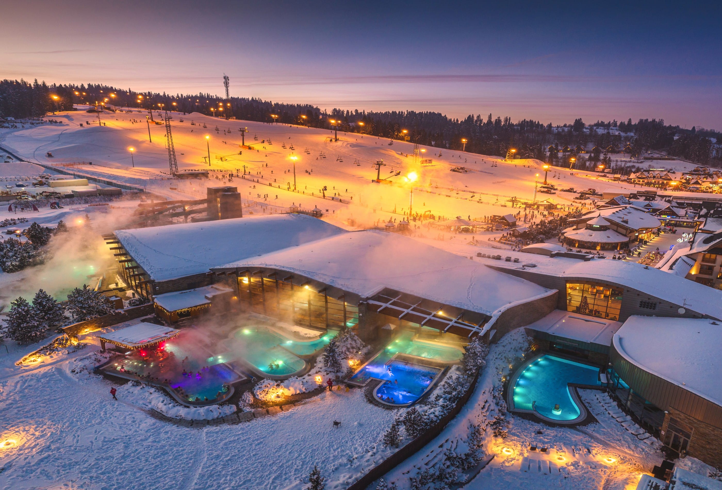 Terma Bania outdoor thermal pools illuminated at night in winter with lit ski slope and snow all around