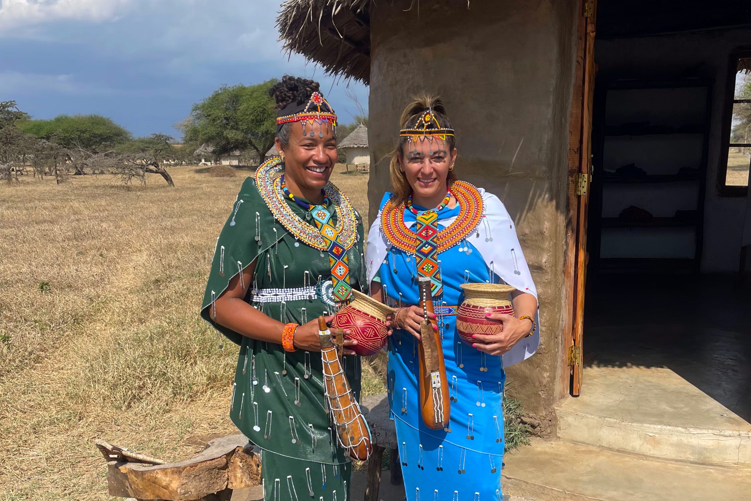 Two women wearing traditional Maasai clothing and beadwork standing outside a hut.