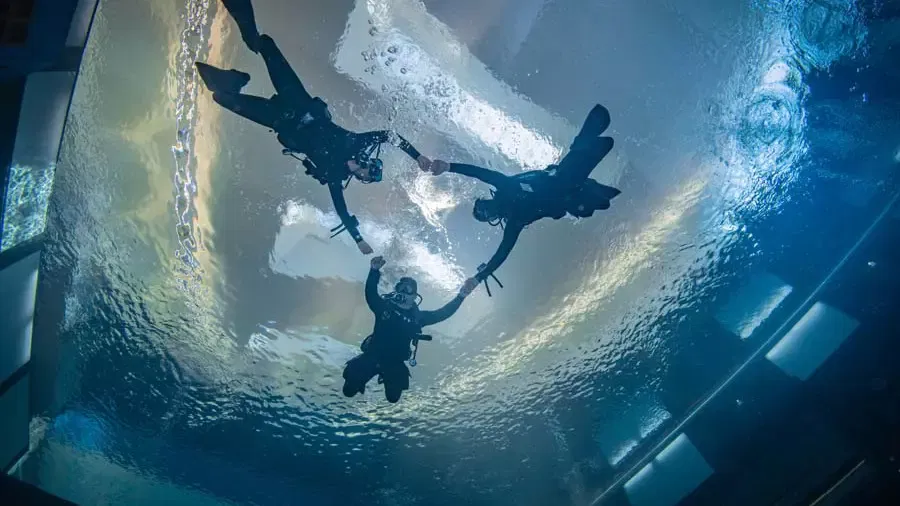 Three scuba divers in full gear hold hands in a circle while floating upward toward the surface at Deep Dive Dubai.